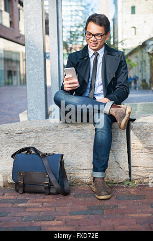Young smiling asian business man looking at mobile phone sitting outside on city street Stock Photo