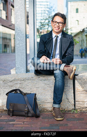 Young smiling asian business man looking at camera holding mobile phone sitting outside on city street Stock Photo
