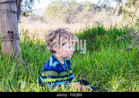 Three year old blonde boy looking into the distance sitting in the long grass on a farm Stock Photo