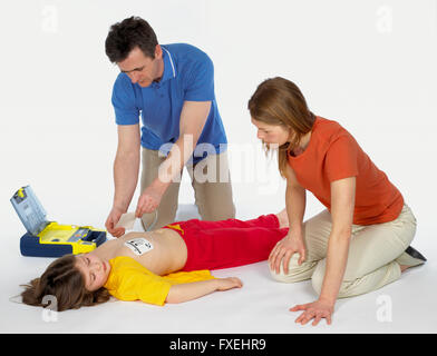 First aid, using paediatric defibrillator, elementary age girl lying on floor unconscious, mature man attaching defibrillator pads to girl's chest, mid adult woman watching Stock Photo