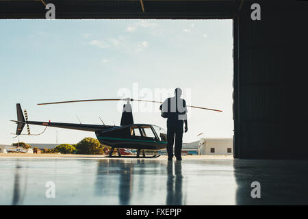 Rear view shot of pilot walking towards helicopter. Pilot and helicopter in a airplane hangar. Stock Photo
