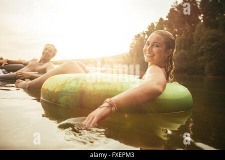 Portrait of happy young woman in lake on inflatable ring with her boyfriends in background. Young couple relaxing in water on a Stock Photo