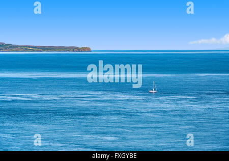 Small sailboat on the Atlantic Ocean near the coast of Northern Ireland with a cliff and a lighthouse Stock Photo
