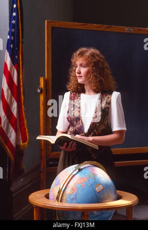 Female red headed teacher in classroom with world globe, American Flag & chalk board Stock Photo