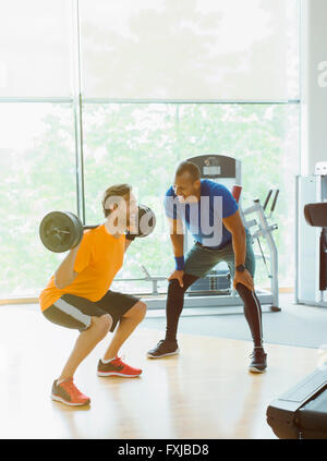 Personal trainer guiding man doing barbell squats at gym Stock Photo
