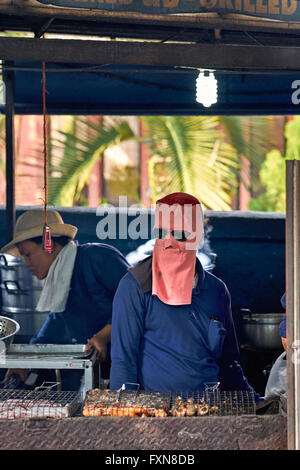 Face mask. Chef cooking at an outdoor market BBQ stall and wearing full face mask to protect against the heat. Thailand S. E. Asia Stock Photo