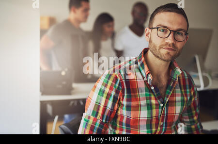 Serious single male business owner wearing multi-colored flannel button shirt with three workers on computer in background
