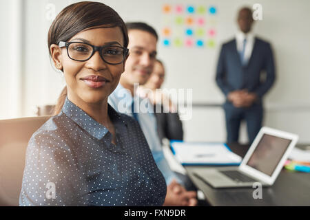 Beautiful cheerful professional woman wearing eyeglasses seated with male co-workers and team leader in conference room at work Stock Photo