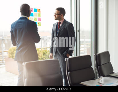 Two business man in stylish suits standing having a discussion in the office looking at a colorful array of sticky memos on the Stock Photo