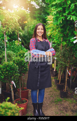 Proud successful confident young nursery owner standing in her greenhouse between the rows of potted plants with folded arms smi Stock Photo