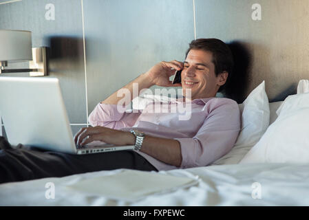Man lying in bed, using cell phone and laptop computer Stock Photo