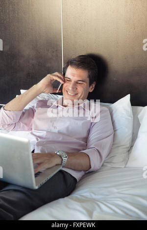 Man using laptop computer and cell phone in bed Stock Photo