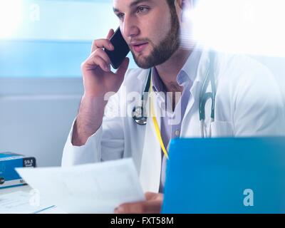 Junior doctor using smartphone whilst reviewing medical records in clinic Stock Photo