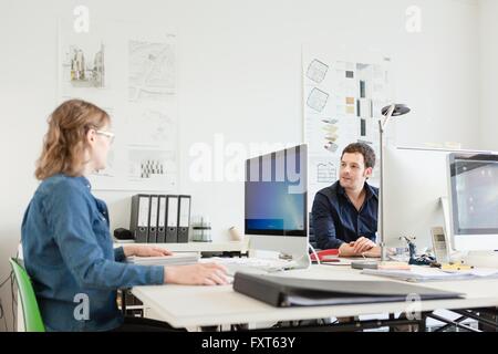 Colleagues sitting at desk in office using computers talking Stock Photo