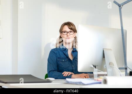 Young woman wearing eyeglasses sitting at desk using computer looking away smiling Stock Photo