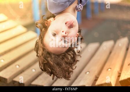 Girl in playground hanging upside down looking at camera sticking out tongue Stock Photo