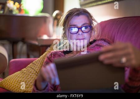 Mature woman wearing eye glasses reclining on sofa using digital tablet Stock Photo