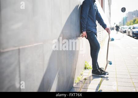 Young male urban skateboarder leaning against sidewalk wall Stock Photo