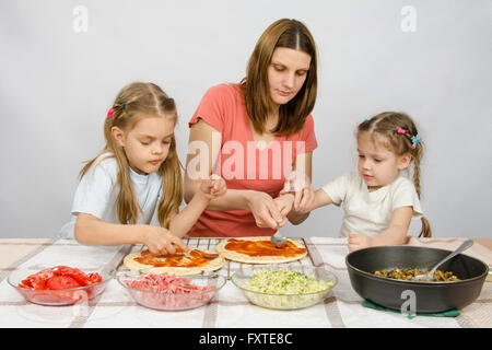 Mom helps younger daughter spread ketchup on a pizza, the eldest daughter, she is preparing a second pizza Stock Photo