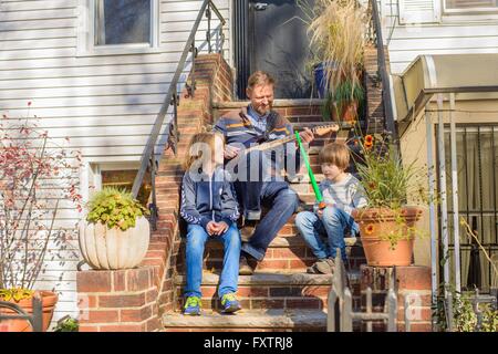 Father and sons sitting together on front steps, father playing guitar Stock Photo