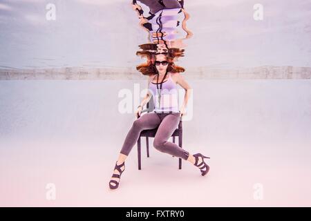 Underwater front view of mid adult woman wearing high heels and sunglasses in swimming pool sitting on chair looking at camera Stock Photo