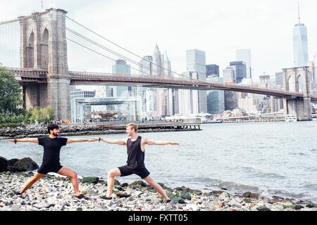 Two men practicing yoga on riverside in front of Brooklyn Bridge, New York, USA Stock Photo