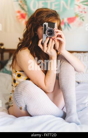 Young woman sitting on bed, looking through SLR camera Stock Photo