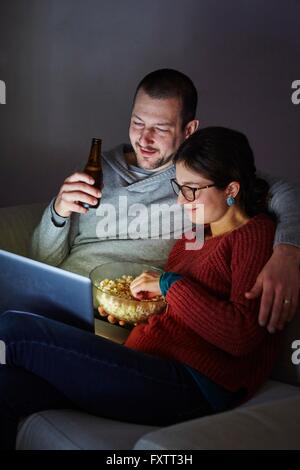 Mid adult couple sitting on sofa in evening eating popcorn and looking at laptop Stock Photo