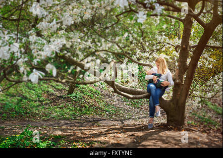 Mom with baby boy sitting on tree branch in the flowered spring garden Stock Photo