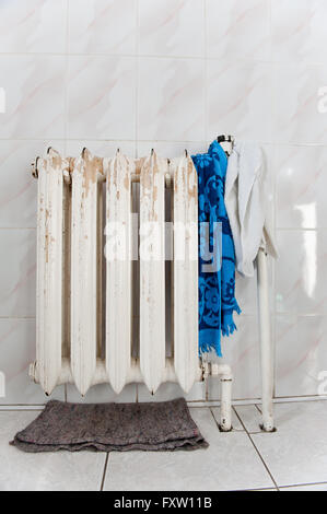 Old cast iron radiator, central heating device with hot water, white paint scratched, few rags drying hanging on it. Stock Photo
