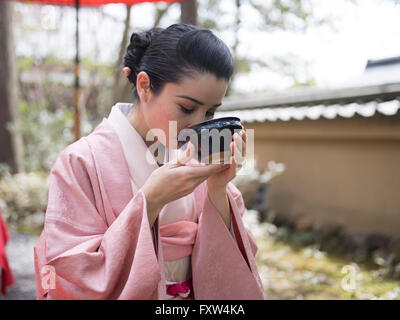 Japanese woman in kimono drinking a bowl of green tea in the gardens of Kinkaku-ji,  a Zen Buddhist temple in Kyoto, Japan. Stock Photo