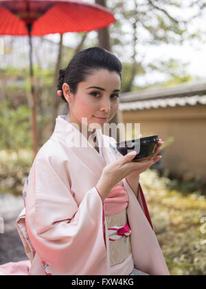 Japanese woman in kimono drinking a bowl of green tea in the gardens of Kinkaku-ji,  a Zen Buddhist temple in Kyoto, Japan. Stock Photo