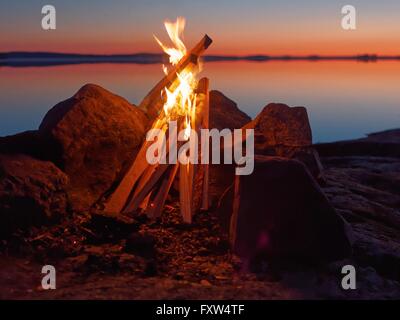 Fire and flames of atmospheric campfire on the beach at night. Still water of the lake on the backgound with warm colors of suns Stock Photo
