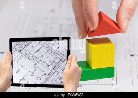 In the bottom left of the photo are hands holding tablet, whose screen contains closeup photo of blueprint of the house . Stock Photo