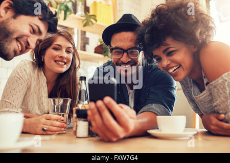 Group of friends sitting together in a cafe looking at mobile phone and smiling. Young guy showing something to his friends on h Stock Photo