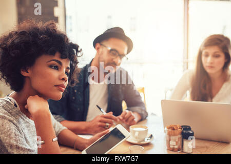 Close up portrait of young african woman with digital tablet and people in background at a cafe table. Young people sitting at a Stock Photo