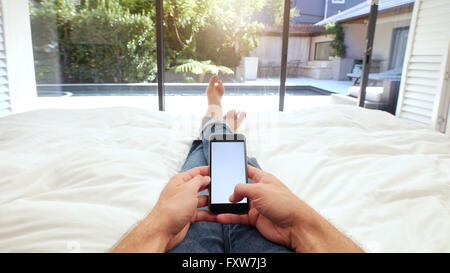 Closeup image of human hand holding mobile phone with blank screen on bed. POV shot of man lying on bed using smart phone. Stock Photo