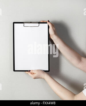 Clipboard with a blank sheet of paper in female hands with a deep shadow on the background of gray wall. Stock Photo