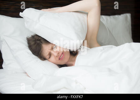 Portrait of young woman lying in bed and covering her ears with pillows from noise. Female model with irritated expression Stock Photo