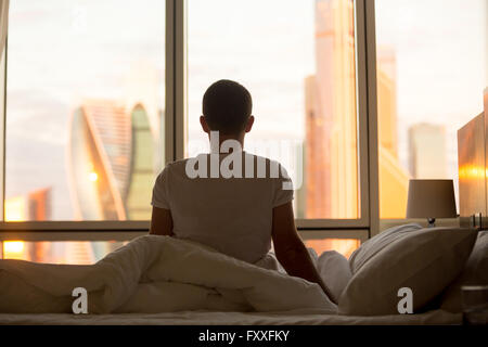 Rear view of young man sitting on unmade bed and looking at dawn city scenery in window after waking up. Handsome casual guy Stock Photo