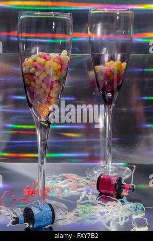 A Studio Photograph of Two Champagne Flutes Containing Sweets and Party Poppers Set Against a Silver Holographic Backdrop Stock Photo