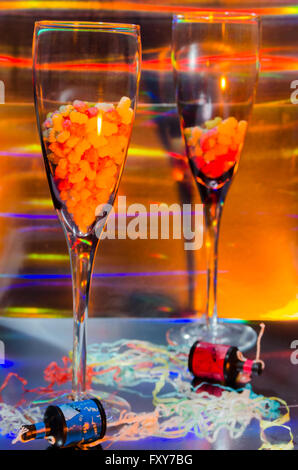 A Studio Photograph of Two Champagne Flutes Containing Sweets and Party Poppers Set Against an Orange Holographic Backdrop Stock Photo