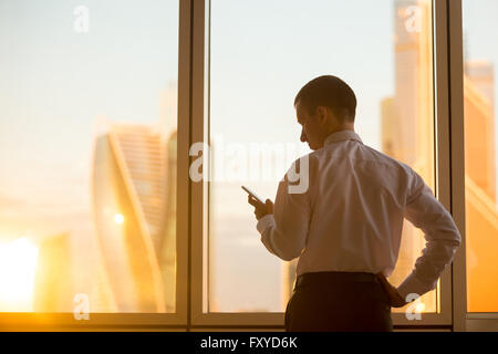 Morning in business office. Rear view of handsome young businessman standing in confident pose and using mobile phone Stock Photo