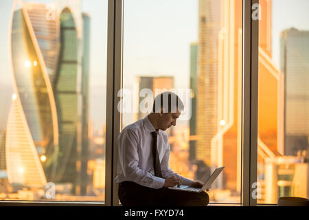 Morning in business office. Handsome young businessman using laptop while sitting in confident pose on panoramic window Stock Photo