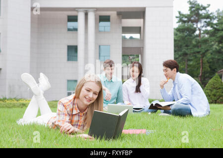 International college student lying on a grass and reading a book and three friends behind her studying together on grass on campus Stock Photo