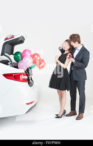 Young man in suit covering woman's eyes to propose to her with present in his hand behind a car with balloons Stock Photo