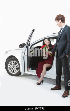 Young smiling woman sitting in oepn car taking a bunch of red roses from a man in suit standing in front of the car Stock Photo