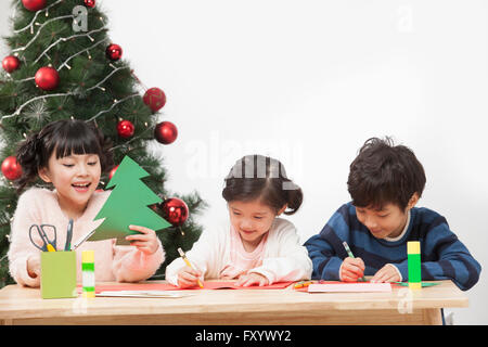 Portrait of three smiling children making paper tree against Christmas tree Stock Photo