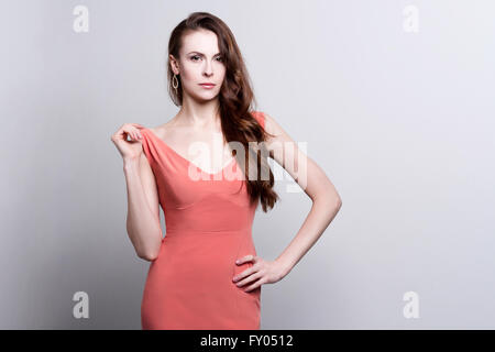 Portrait of a young attractive woman with beautiful long brown hair. She wears a coral dress and golden earrings. She touches he Stock Photo