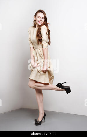 Attractive young woman in golden dress smiles and stands in fashion pose, she weares black high heels. She has long brown hair.